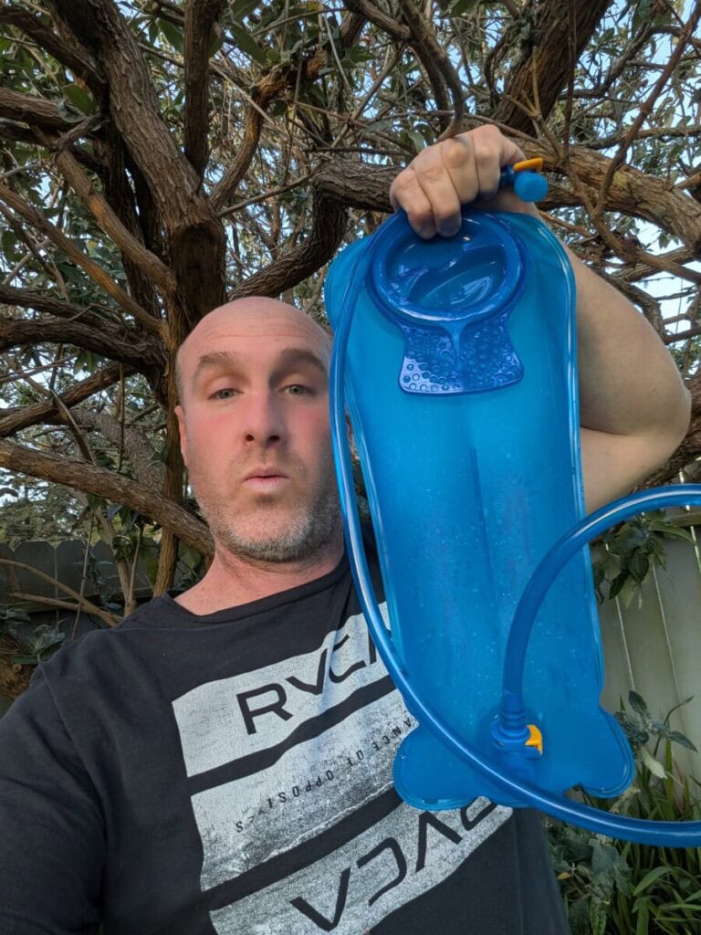 Me holding up a 3L bladder that holds water, a tree in the background. Some wonder how to save money on water while traveling.